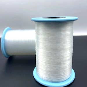 May include: Two spools of white thread. The thread is tightly wound around a light blue spool. The thread appears to be a translucent, fine fiber. The spools are set against a neutral background.