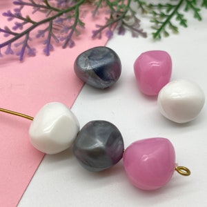 K&ouml;nnte beinhalten: Eine Perlenkette mit einer Mischung aus Farben und Texturen. Die Perlen sind wei&szlig;, rosa und grau mit einer metallischen Oberfl&auml;che. Die Perlen sind auf einem goldenen Draht aufgef&auml;delt.