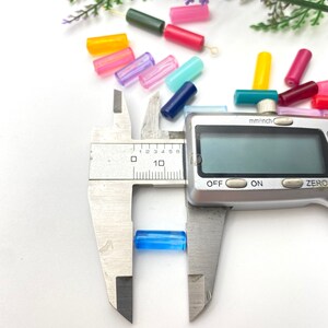 May include: A blue glass bead is being measured with a digital caliper. The caliper reads 0.6 cm. The bead is one of many colorful glass beads scattered on a white surface.