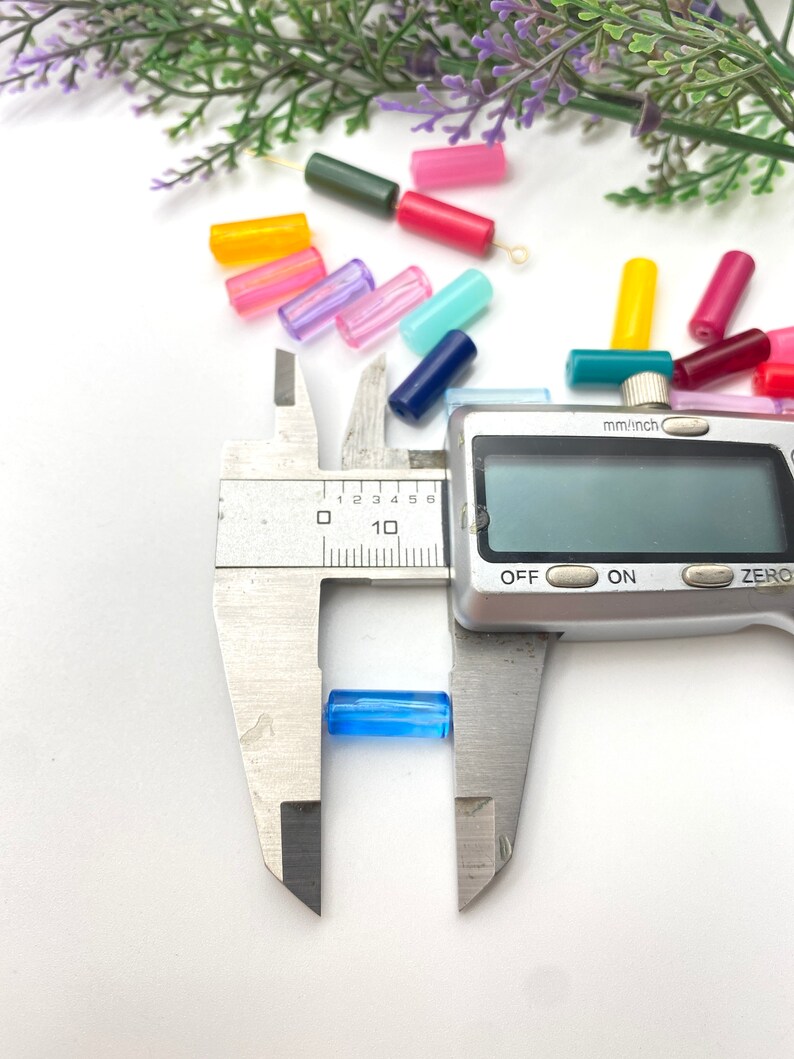May include: A blue glass bead is being measured with a digital caliper. The caliper reads 0.6 cm. The bead is one of many colorful glass beads scattered on a white surface.
