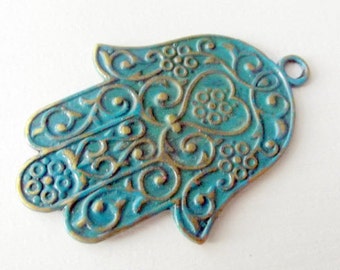 May include: A turquoise blue hamsa hand charm with gold accents and intricate floral designs. The charm has a loop at the top for hanging.