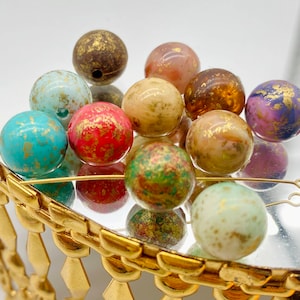 May include: A collection of round, colourful glass beads with gold flecks. The beads are arranged in a pile on a gold-coloured surface.