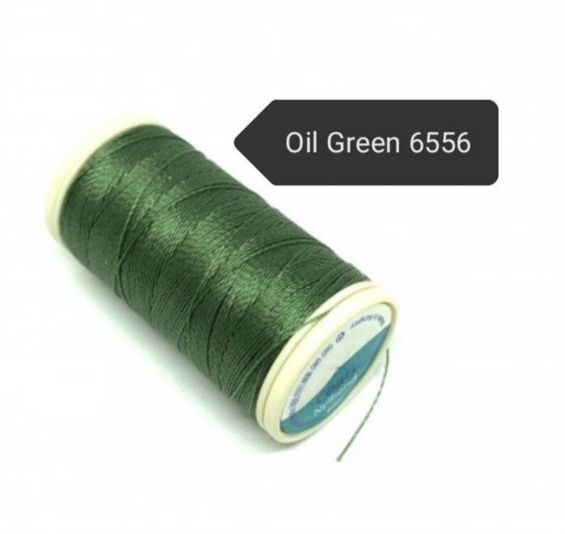 May include: A spool of oil green thread, color code 6556, is displayed against a white background. The thread is tightly wound around a white and beige spool. The image is well-lit, highlighting the thread's texture and color.