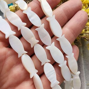 Może przedstawiać: Zbliżenie na sznury białych koralików w kształcie ryb. Koraliki mają perłowy połysk i są nawleczone. Są trzymane w dłoni, z żółtymi kwiatami w tle. Nadaje się do tworzenia biżuterii.