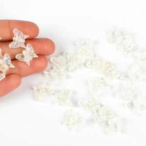 May include: A collection of small, iridescent butterfly-shaped embellishments. Some butterflies are held in a hand, while others are scattered on a white surface. The butterflies have a translucent, glittery appearance.