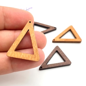 Può includere: Elementi artigianali a forma di triangolo in legno in varie tonalità di marrone. I triangoli aperti hanno un piccolo foro nella parte superiore per l'infilatura. I pezzi sono tenuti in mano su sfondo bianco, ideali per la creazione di gioielli o altri lavori artigianali.