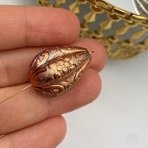 Puede incluir: Una cuenta de color cobre con un intrincado diseño floral. La cuenta es de forma ovalada y tiene un pequeño agujero en la parte superior para enhebrar.