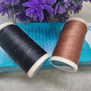 May include: Two spools of Nylbond thread, one black and one brown, sit on a blue box with text. The spools have white rims and are next to purple flowers. The text on the box includes "Extra Strong Nylon".