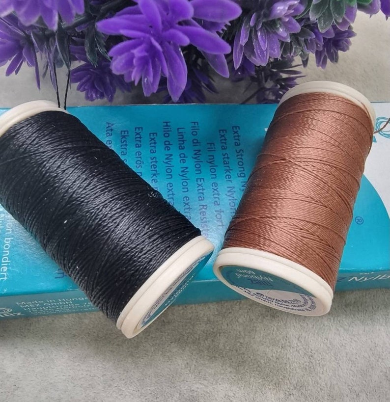 May include: Two spools of Nylbond thread, one black and one brown, sit on a blue box with text. The spools have white rims and are next to purple flowers. The text on the box includes "Extra Strong Nylon".