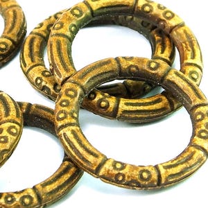 May include: Close-up of several decorative rings. The rings are a light brown color with darker brown accents. The rings have a textured surface with a repeating pattern of circles and lines. The rings are arranged in a loose pile.