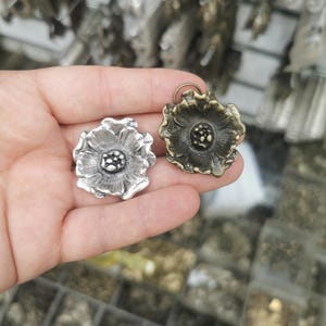 Puede incluir: Dos colgantes con forma de flor, uno plateado y otro bronce, se sostienen en una mano. Los colgantes tienen diseños de pétalos detallados y un centro texturizado. El fondo muestra otros artículos similares.
