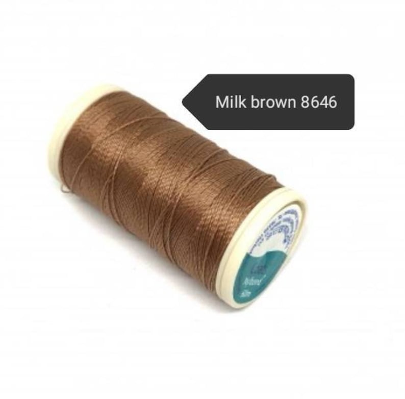 May include: A spool of milk brown thread, color code 8646, is shown. The thread is tightly wound around a white and teal spool. The thread is a light brown color.