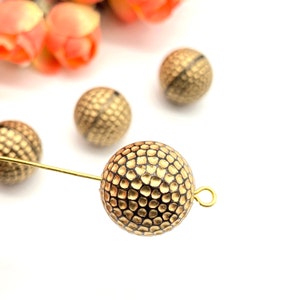 Może przedstawiać: Okrągłe koraliki w kolorze złotym i czarnym o fakturowanej powierzchni. Koraliki mają małą pętelkę do przymocowania do sznurka lub łańcuszka.