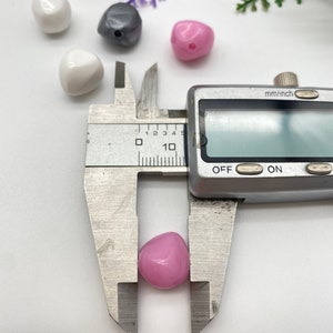 K&ouml;nnte beinhalten: Nahaufnahme einer rosafarbenen herzf&ouml;rmigen Plastikperle, die mit einer Schieblehre gemessen wird. Die Schieblehre zeigt 1,0 cm an.