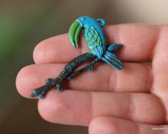 May include: A blue and green toucan bird charm with a long beak, perched on a branch. The charm is made of metal and has a rustic finish.