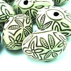 Pode incluir: Um grupo de contas de cerâmica ovais com um esmalte branco e desenhos florais marrons. As contas têm um furo no centro para serem enfiadas.