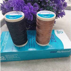 May include: Two spools of MEZ Nylbond thread, one black and one bronze, sit atop a teal box. The spools are labeled with the brand name and 60m of thread. The box has text in multiple languages. Purple flowers are in the background.