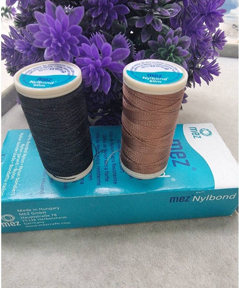 May include: Two spools of MEZ Nylbond thread, one black and one bronze, sit atop a teal box. The spools are labeled with the brand name and 60m of thread. The box has text in multiple languages. Purple flowers are in the background.