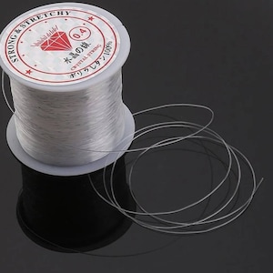 Könnte beinhalten: Eine Spule aus klarem elastischem Garn mit einem Etikett mit der Aufschrift "Strong & Stretchy 0.4 水田の線 Crystal String オリカレタ 100%"