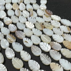 Könnte beinhalten: Nahaufnahme einer Sammlung von blattförmigen Perlen. Die Perlen sind überwiegend weiß mit subtilen Variationen von Beige und Hellbraun. Jede Perle hat detaillierte Adern, was auf ein natürliches Material hindeutet. Die Anordnung deutet darauf hin, dass sie für die Schmuckherstellung bestimmt sind.