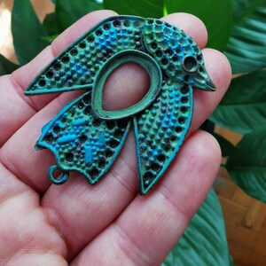 Może przedstawiać: Metalowy wisiork w kształcie ptaka w kolorze turkusowym i zielonym, z fakturowaną powierzchnią i małą pętelką do przymocowania do naszyjnika lub bransoletki.