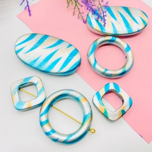 May include: Five ceramic beads with a blue and silver zebra print design. The beads are in various shapes, including oval, round, and square. Some of the beads have holes for threading.