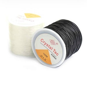 Pode incluir: Dois rolos de fio elástico preto e branco, cada um com a etiqueta "Crystal Tec" com "100M" e "Ø1.0" impressos na etiqueta.