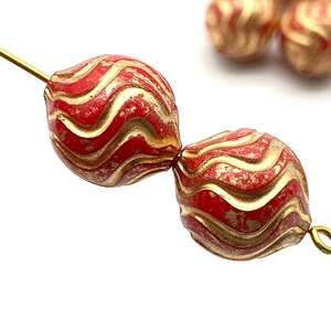 May include: Two red and gold glass beads with a wavy pattern. The beads are strung on a gold-colored wire and have a loop at the end. Additional beads are visible in the background, suggesting a jewelry-making theme.