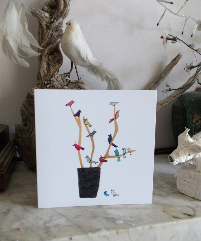 May include: A white card with a drawing of a tree with many colorful birds perched on the branches. The tree is in a gray pot. The card is on a white marble surface.