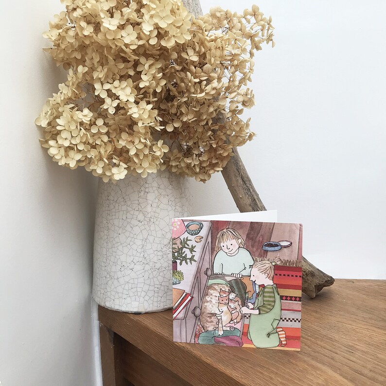 Pu&ograve; includere: Un biglietto d'auguri con un'illustrazione di due bambini che guardano un gruppo di gattini. La carta ha un bordo a strisce rosso, bianco e blu.