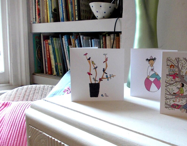 May include: Three greeting cards with different illustrations. The first card features a tree with birds, the second card features a person sitting on a beach ball, and the third card features a tree with people sitting on branches.
