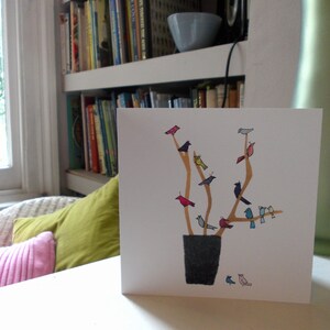 May include: A white card with a gold and brown tree with colorful birds perched on the branches. The tree is in a black pot. The card is on a white surface.