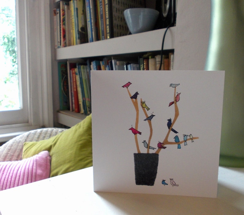 May include: A white card with a gold and brown tree with colorful birds perched on the branches. The tree is in a black pot. The card is on a white surface.