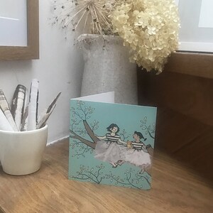 Pu&ograve; includere: Un biglietto d'auguri con uno sfondo blu chiaro che presenta due ragazze sedute su un ramo d'albero con un piccolo gatto. Le ragazze indossano camicie a righe e gonne bianche. Il ramo dell'albero &egrave; circondato da foglie verdi.