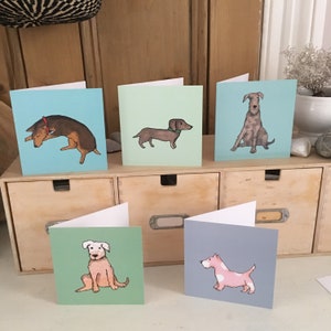 May include: Five greeting cards featuring illustrations of various dog breeds. The cards are displayed in a row on a wooden shelf. The dogs are depicted in a simple, cartoon style. Each card is a different colour.