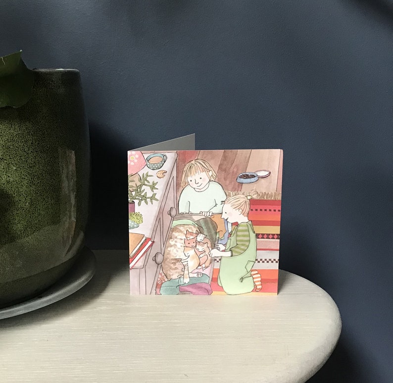 Pu&ograve; includere: Un biglietto d'auguri con un'illustrazione di due bambini che guardano dentro un cassetto di una cassettiera con tre gattini dentro. Il cassetto &egrave; aperto e i bambini sorridono. La carta ha uno sfondo a righe colorato.