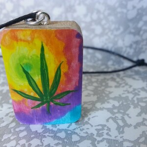 May include: A wooden pendant with a tie-dye background and a green marijuana leaf painted on it. The pendant is attached to a black cord.
