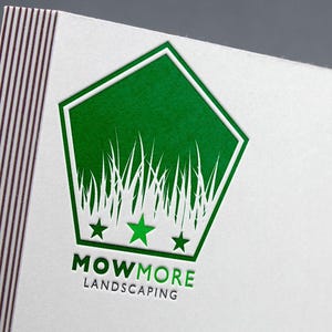 A close-up of a white business card with a green foil-stamped pentagonal logo featuring grass, three stars, and "MOWMORE LANDSCAPING" text.