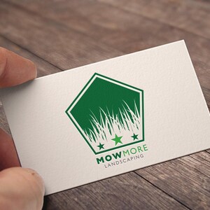 A hand holding a white business card with a green pentagonal logo featuring grass, three stars, and "MOWMORE LANDSCAPING" text, against a wooden background.