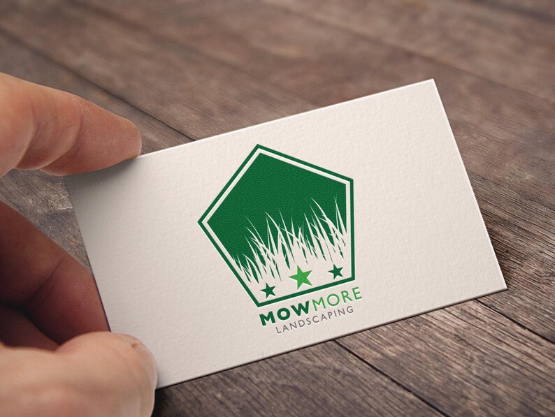 A hand holding a white business card with a green pentagonal logo featuring grass, three stars, and "MOWMORE LANDSCAPING" text, against a wooden background.