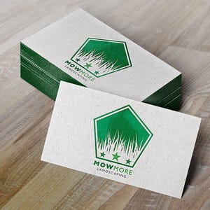 A stack of white business cards with a green pentagonal logo featuring grass, three stars, and "MOWMORE LANDSCAPING" text, next to a single card on a wooden surface.