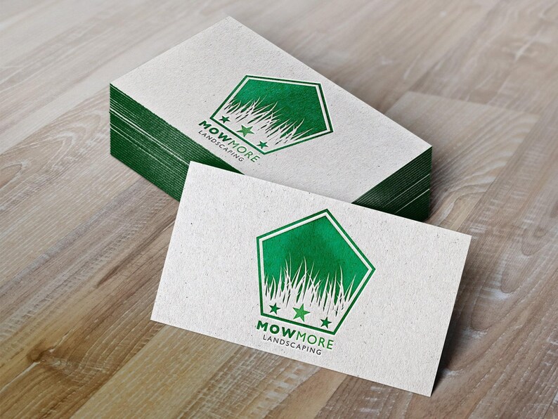 A stack of white business cards with a green pentagonal logo featuring grass, three stars, and "MOWMORE LANDSCAPING" text, next to a single card on a wooden surface.