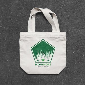 A small white canvas tote bag with a green pentagonal logo featuring grass, three stars, and "MOWMORE LANDSCAPING" text, on a dark gray textured surface.