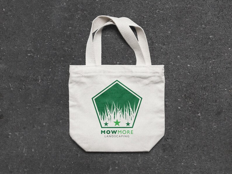 A small white canvas tote bag with a green pentagonal logo featuring grass, three stars, and "MOWMORE LANDSCAPING" text, on a dark gray textured surface.