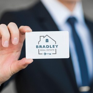 A hand in a dark suit holds a white business card with a gray and blue logo of a house outline, "BRADLEY REAL ESTATE," and a vintage key.