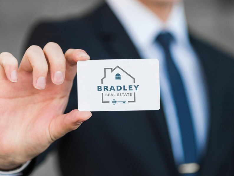 A hand in a dark suit holds a white business card with a gray and blue logo of a house outline, "BRADLEY REAL ESTATE," and a vintage key.