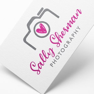 May include: A white business card with a gray camera icon and a pink heart inside. The text "Sally Sherman Photography" is written in pink cursive.