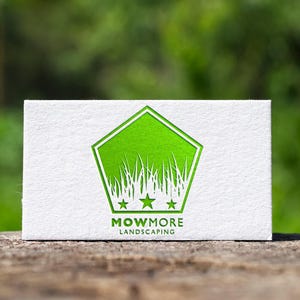 A white business card with a green pentagonal logo featuring grass, three stars, and "MOWMORE LANDSCAPING" text, resting on a wooden surface with a blurred green background.