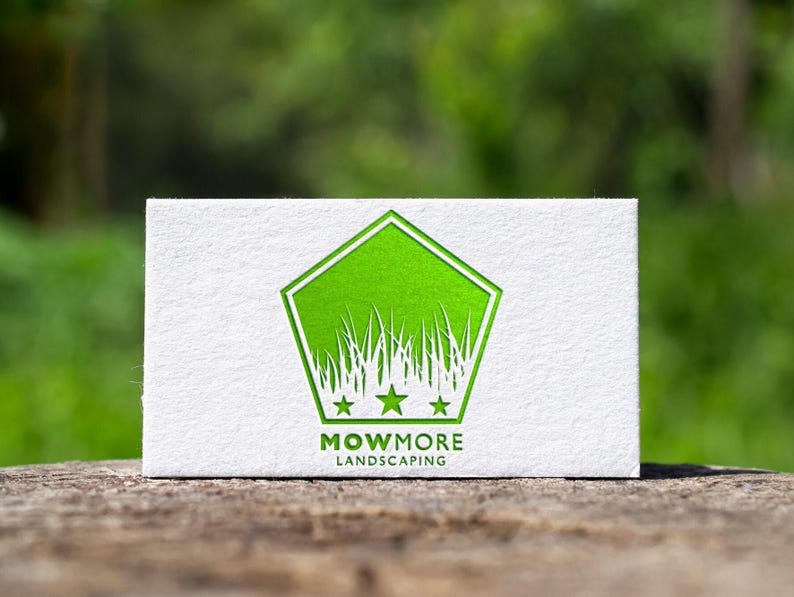 A white business card with a green pentagonal logo featuring grass, three stars, and "MOWMORE LANDSCAPING" text, resting on a wooden surface with a blurred green background.