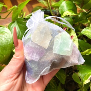 May include: A small white organza bag filled with colorful crystals. The crystals are various shades of green, purple, and clear.